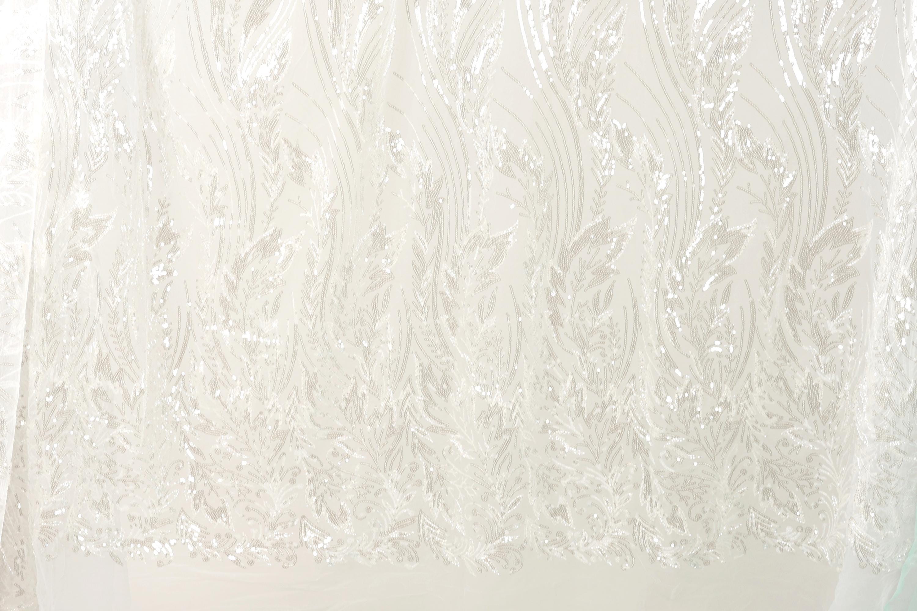 The image is a close-up of a white fabric with a repeating pattern of white leaves or flowers. The fabric appears to be made of a sheer or sheer-like material, which gives it a delicate and airy appearance. The pattern is intricate and detailed, with the leaves or flowers appearing to be made of sequins or glitter, adding a touch of sparkle to the fabric. The image does not contain any text or other discernible objects, and the focus is solely on the fabric and its pattern.
