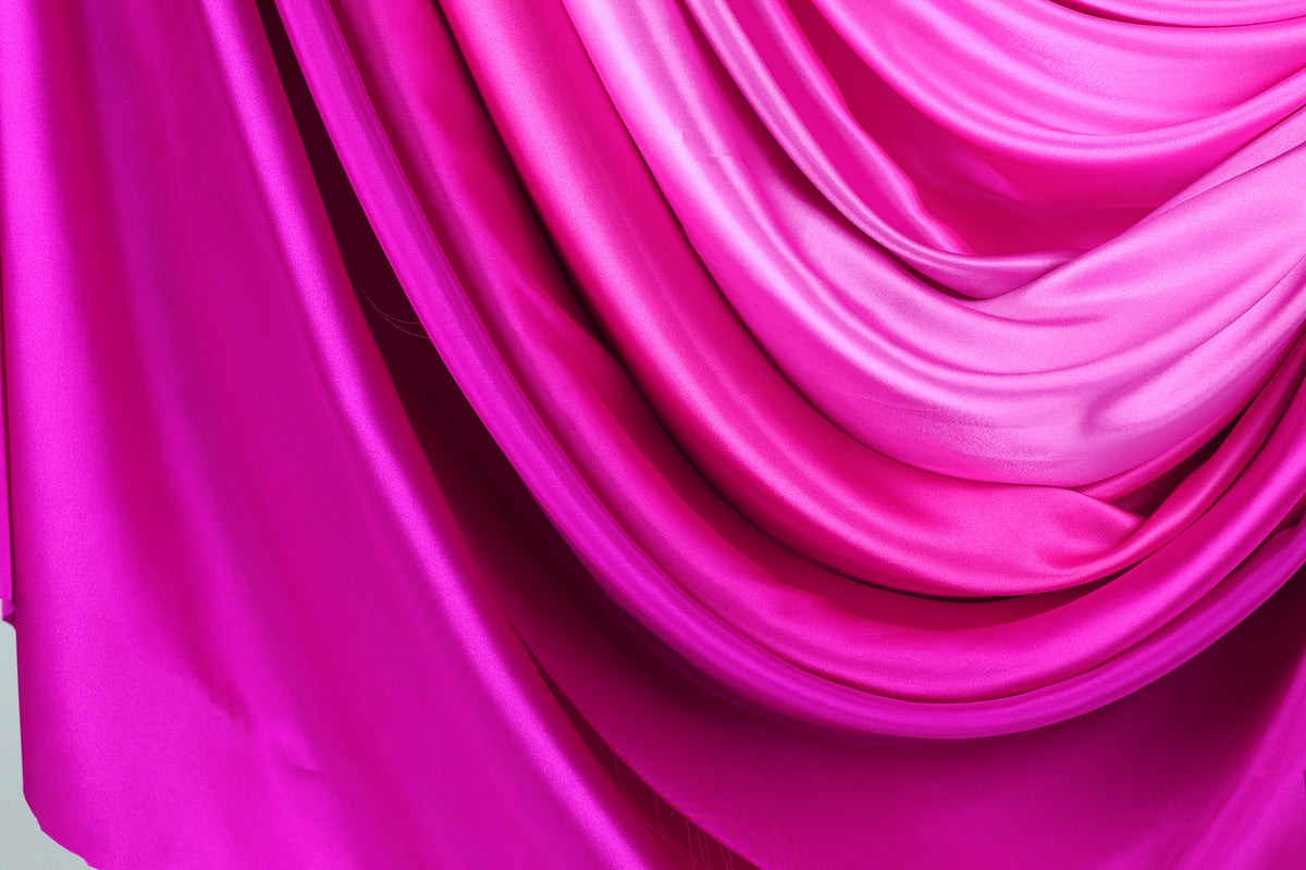 a close-up view of a vibrant, flowing fabric with a gradient of pink and purple colors. The fabric appears to be made of silk or satin, and it is draped in a way that creates a sense of movement and elegance. The colors are rich and intense, with the pink and purple hues dominating the fabric, while the fabric itself has a smooth and shiny texture.
