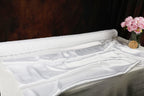 a white satin fabric on a table, with a vase of pink flowers placed next to it.