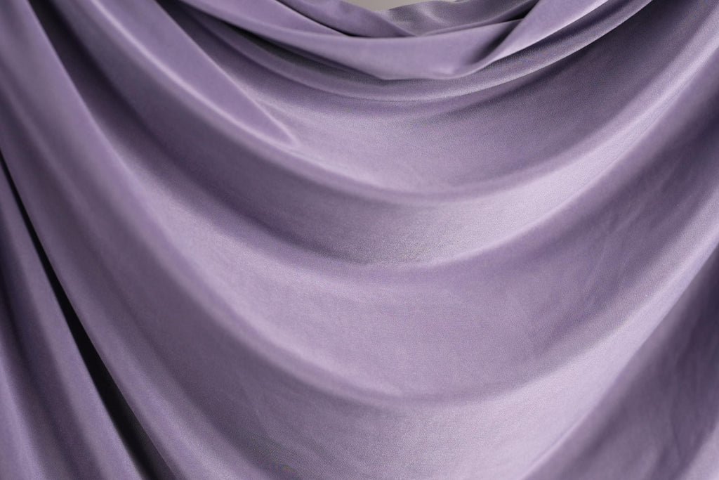 a close-up view of a soft, flowing purple fabric, possibly a curtain or garment. The fabric has a smooth, shiny texture and is draped in a way that creates a sense of movement and depth.